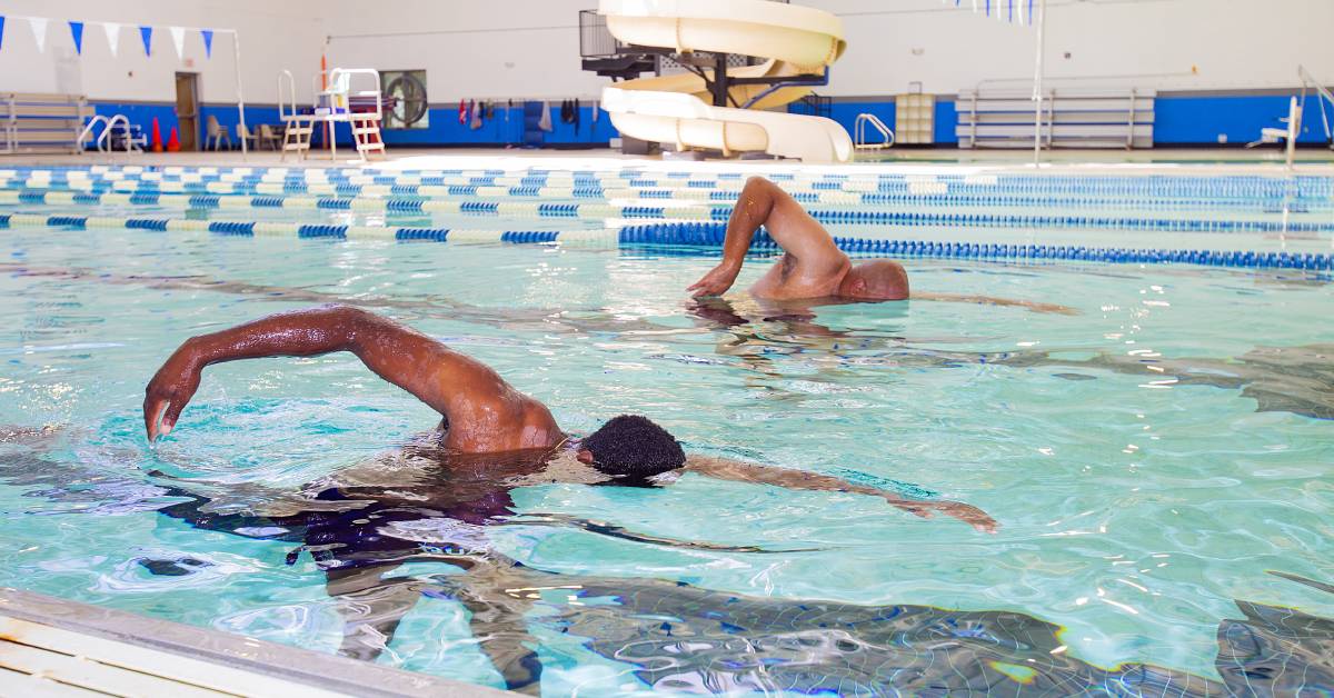 Two people swimming freestyle in an indoor pool with lanes and a water slide in the background.