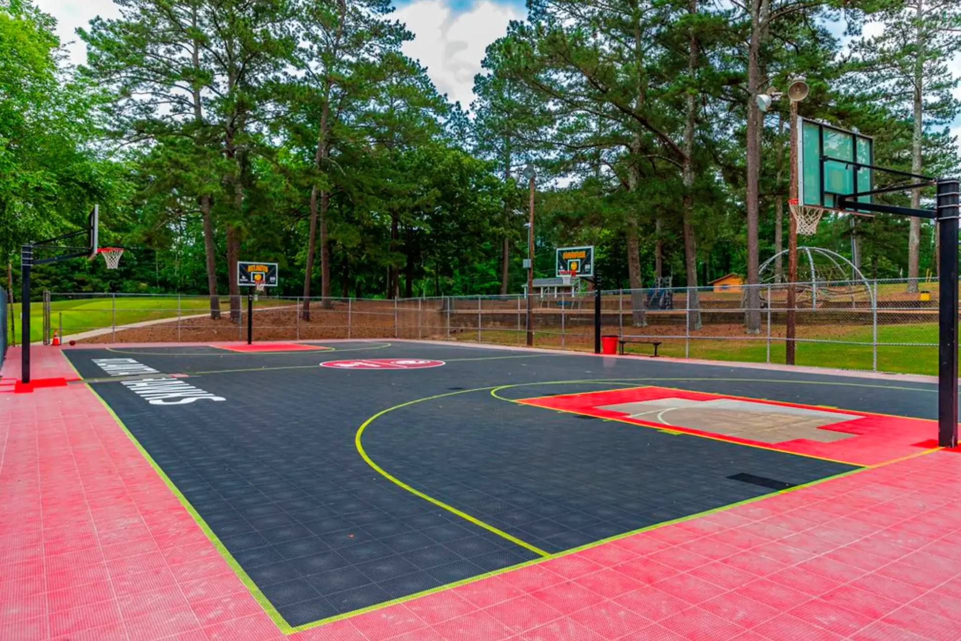 A vibrant outdoor basketball court surrounded by tall trees, featuring multiple hoops, and a colorful playing surface