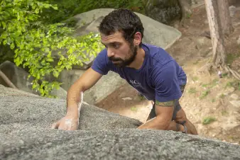 A climber grips a rocky surface, chalked hands poised for ascent amidst lush green foliage and natural surroundings