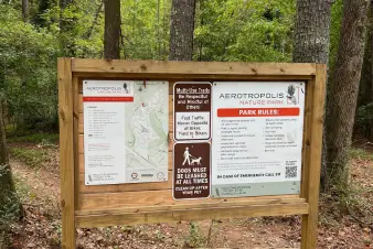 A wooden sign at Aerotropolis Nature Park detailing park rules, trail usage, and pet policies, surrounded by lush trees