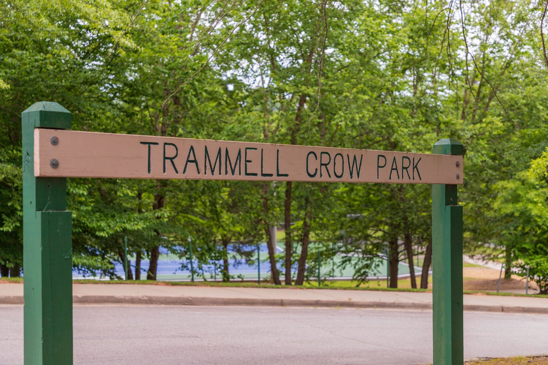 Wooden sign for Trammell Crow Park amidst lush greenery, with tennis courts visible in the background